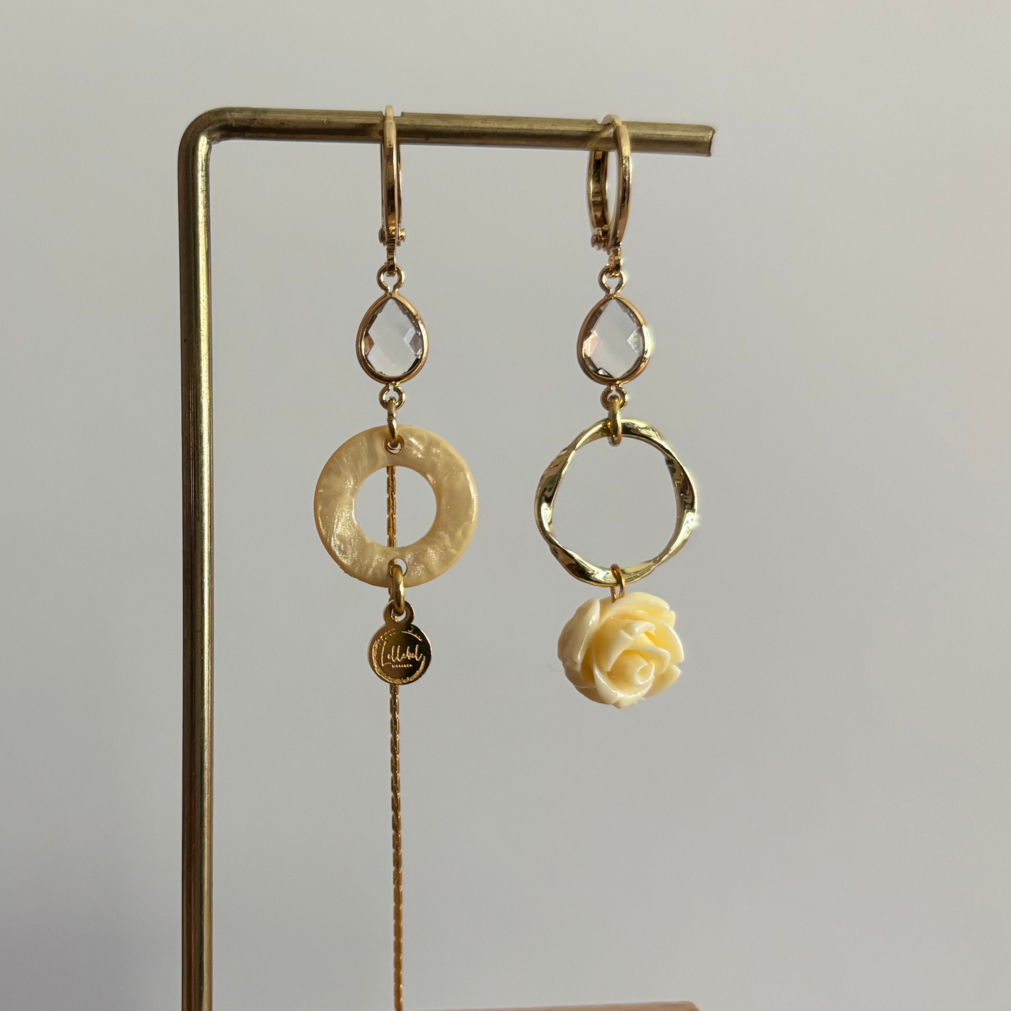 Asymmetrische goudkleurige oorbellen Jolijn aan een rekje. Beide oorbellen hebben een heldere kristaldruppel; links hangt een crèmekleurige ring met een lang kettinkje en rechts een organische gouden cirkel met een crèmekleurig roosje.