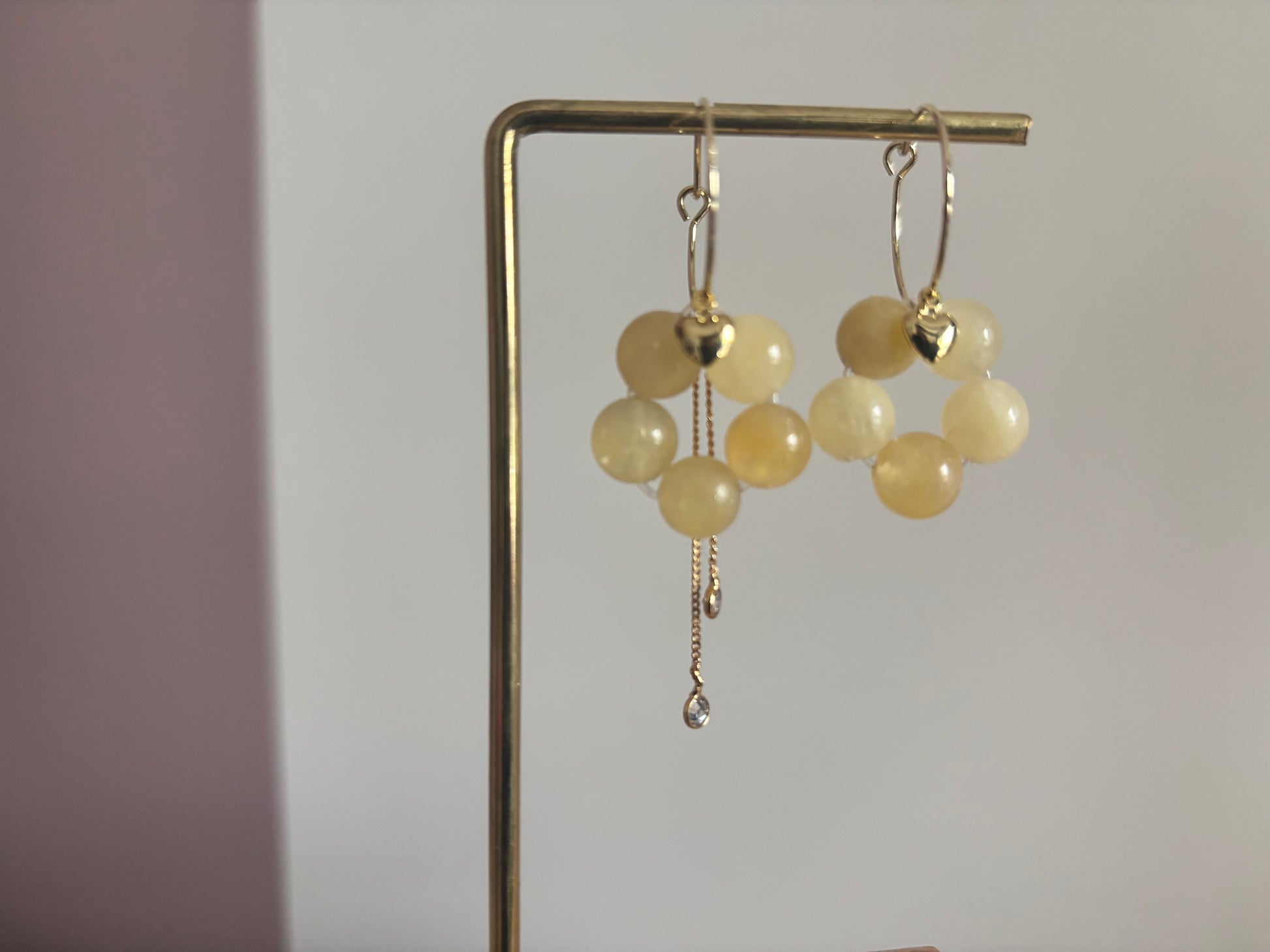 Gouden oorbellen [Naam] met botergele kralen in bloemvorm, voorzien van een hartje en een asymmetrisch kettinkje.