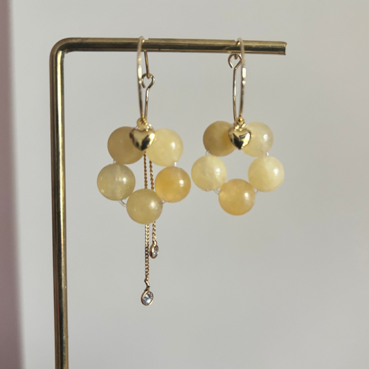 Gouden oorbellen [Naam] met botergele kralen in bloemvorm, voorzien van een hartje en een asymmetrisch kettinkje.