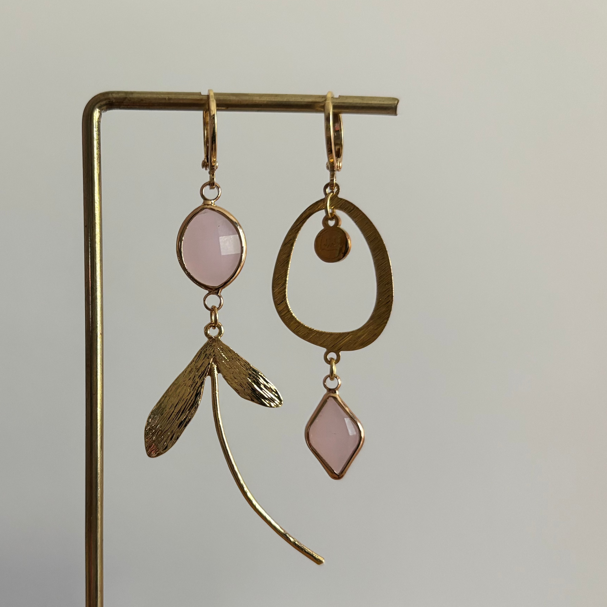Asymmetrische 14k vergulde oorbellen Kate Melkroze aan een rekje. Links een ronde melkroze steen met een botanische bladhanger; rechts een grote open ring met een hangende, ruitvormige melkroze steen.