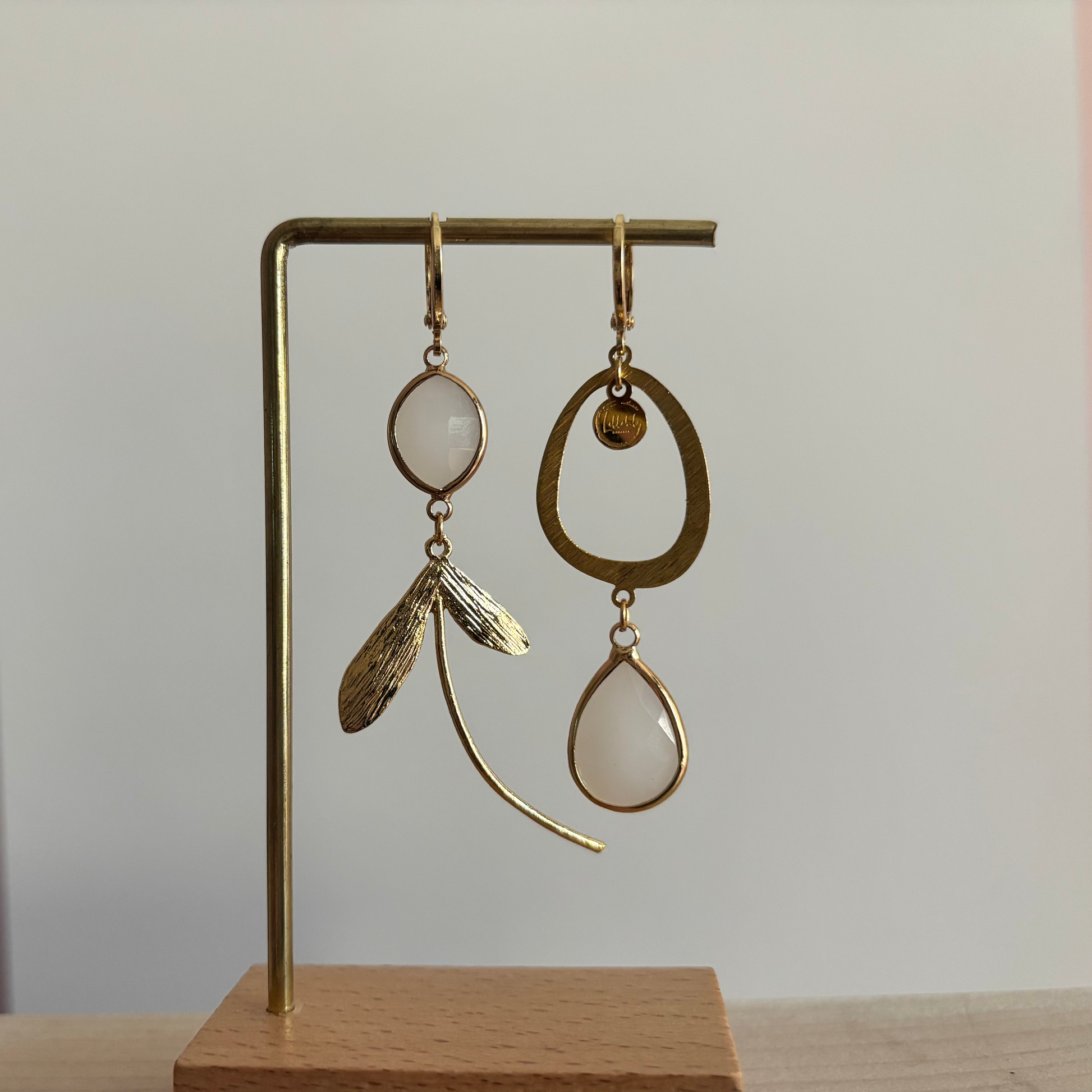 Asymmetrische 14k vergulde oorbellen Kate Melkwit aan een rekje. Links een ovale melkwitte steen met een botanische bladhanger; rechts een grote open ring met een hangende melkwitte kristaldruppel.