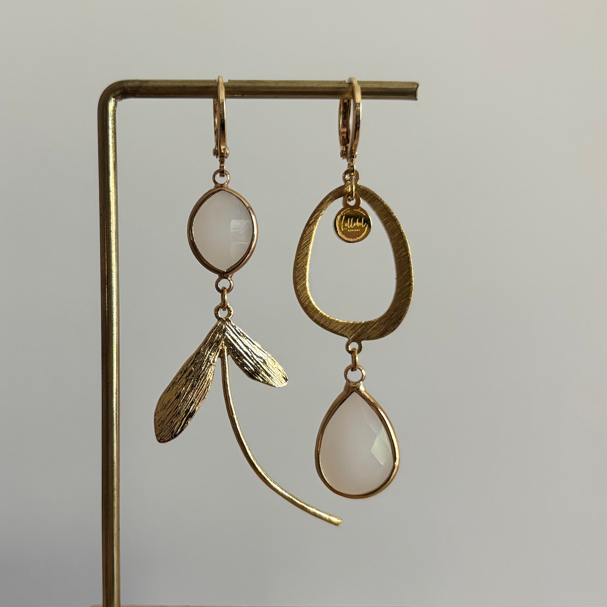 Asymmetrische 14k vergulde oorbellen Kate Melkwit aan een rekje. Links een ovale melkwitte steen met een botanische bladhanger; rechts een grote open ring met een hangende melkwitte kristaldruppel.