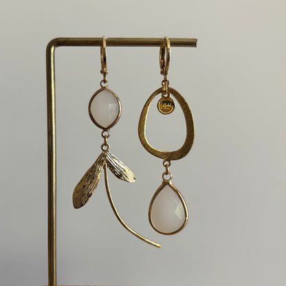 Asymmetrische 14k vergulde oorbellen Kate Melkwit aan een rekje. Links een ovale melkwitte steen met een botanische bladhanger; rechts een grote open ring met een hangende melkwitte kristaldruppel.