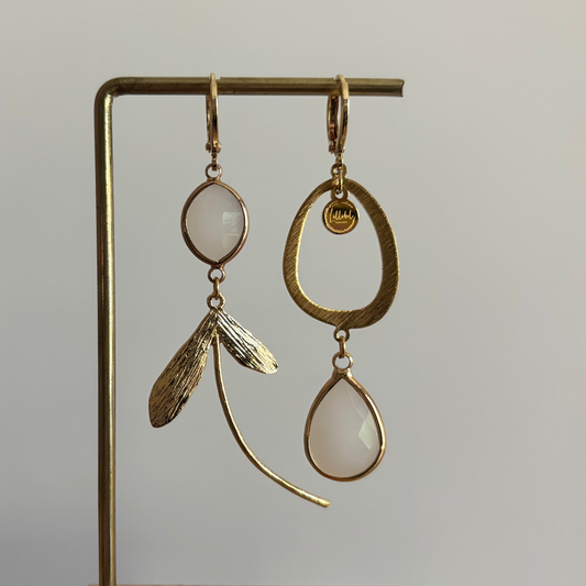 Asymmetrische 14k vergulde oorbellen Kate Melkwit aan een rekje. Links een ovale melkwitte steen met een botanische bladhanger; rechts een grote open ring met een hangende melkwitte kristaldruppel.