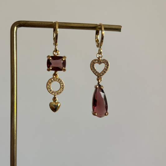 "Gouden mismatched oorbellen set Kelly Burgundy met bordeauxrode glasstenen. De linker oorbel heeft een rechthoekige paarsrode steen, een bewerkt rondje en een goud hartje. De rechter oorbel heeft een opengewerkt strass-hartje en een traanvormige bordeauxrode steen.