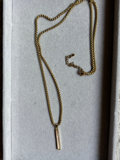Lange goudkleurige ketting Nanet in een grijs sieradendoosje, voorzien van een dunne, ronde schakelketting en een rechthoekige hanger van wit parelmoer schelp.