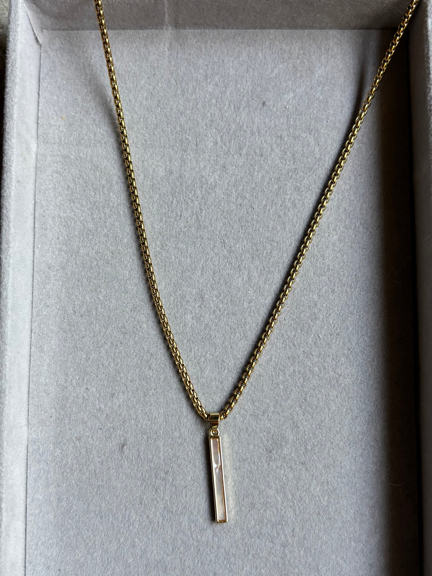 Lange goudkleurige ketting Nanet in een grijs sieradendoosje, voorzien van een dunne, ronde schakelketting en een rechthoekige hanger van wit parelmoer schelp.