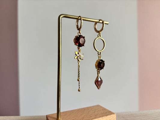 Goudkleurige asymmetrische hangoorbellen Kiara Burgundy met ronde en ruitvormige burgundy kristallen, zirkonia bloemetje en een gouden cirkel.
