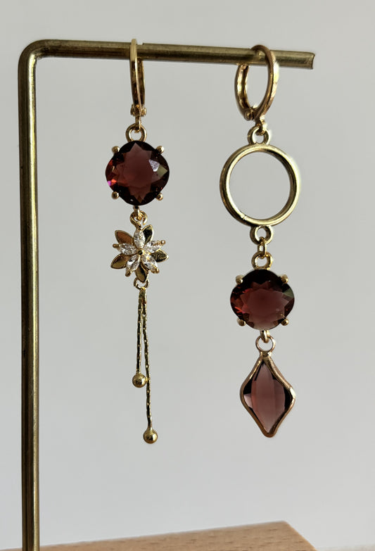 Goudkleurige asymmetrische hangoorbellen Kiara Burgundy met ronde en ruitvormige burgundy kristallen, zirkonia bloemetje en een gouden cirkel.