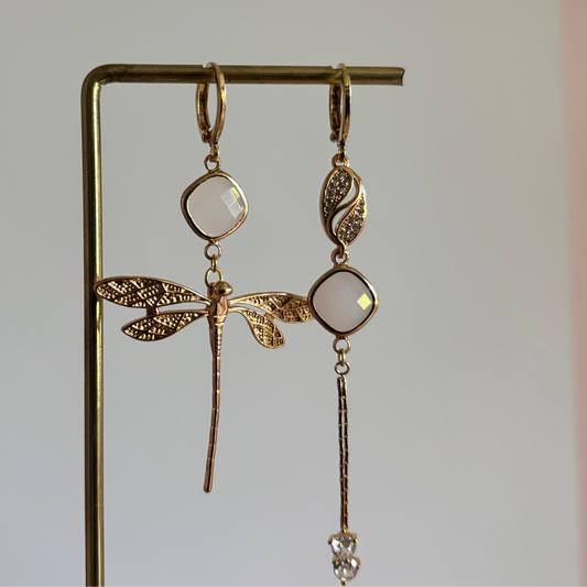 Set asymmetrische goudkleurige oorbellen aan een display. Links een hanger met strass, een melkwitte steen en een fijn kettingkje met steentjes. Rechts een melkwitte steen met daaronder een goudkleurige libelle.