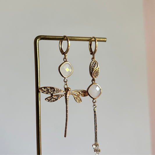 Set asymmetrische goudkleurige oorbellen aan een display. Links een hanger met strass, een melkwitte steen en een fijn kettingkje met steentjes. Rechts een melkwitte steen met daaronder een goudkleurige libelle.