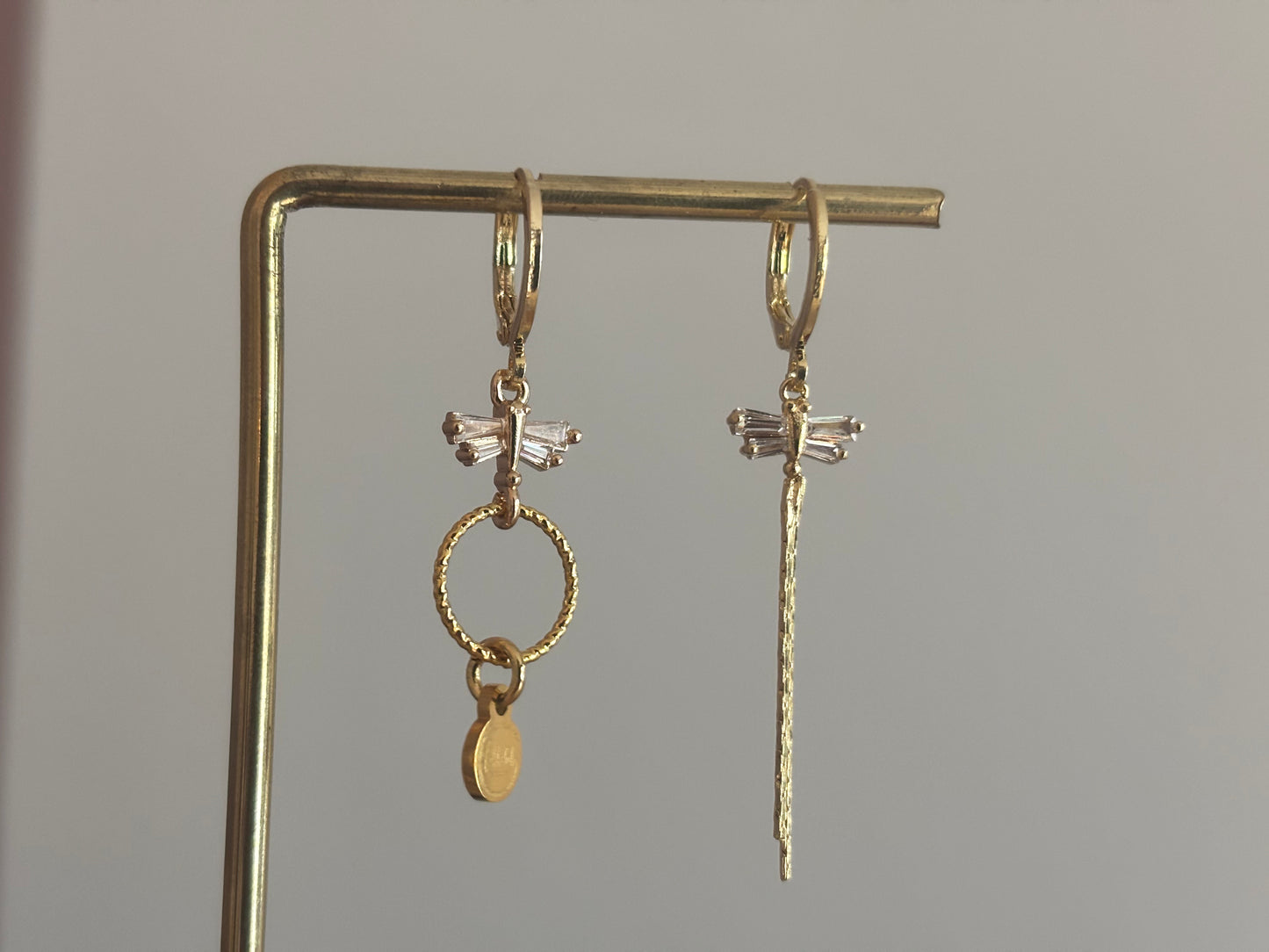 Asymmetrische oorbellen met zirkonia libelles, één met strengetje en één met minimalistische hanger en Lellebel-tag, circa 4 cm lang.