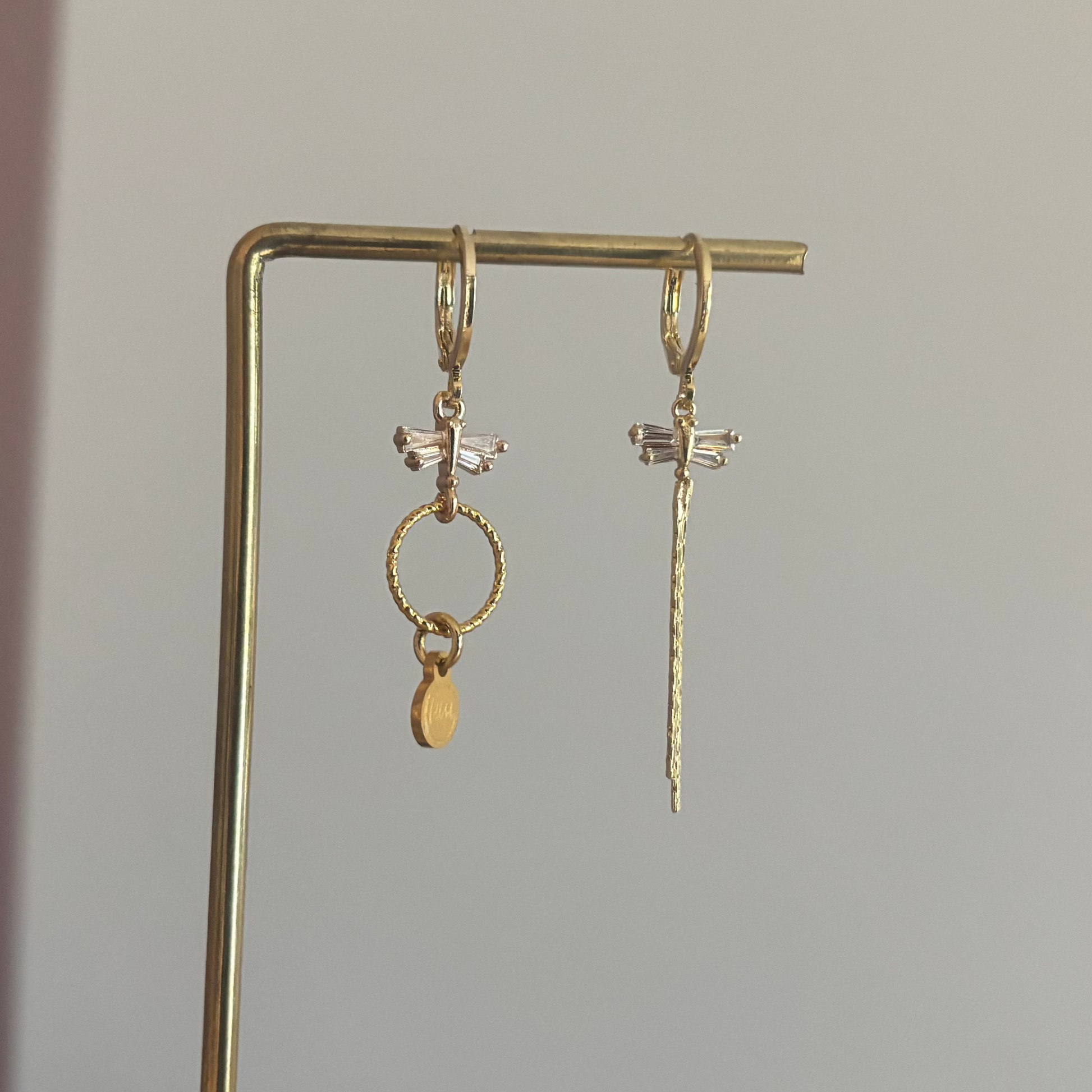 Asymmetrische oorbellen met zirkonia libelles, één met strengetje en één met minimalistische hanger en Lellebel-tag, circa 4 cm lang.