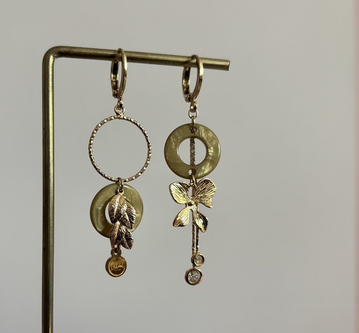 Twee verschillende goudkleurige oorbellen Lieke op een standaard: links een ronde ring met een parelmoerachtige schijf en blaadjes, rechts een goudkleurige strik met hangende strass-steentjes.