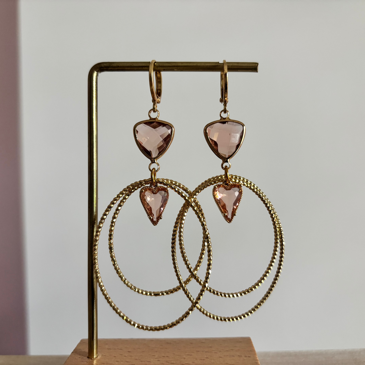 Symmetrische goudkleurige oorbellen Liene aan een rekje, opgebouwd uit een driehoekige champagnekleurige glasbedel, twee robuuste ovale ringen en een klein champagnekleurig hartje als verbinding.