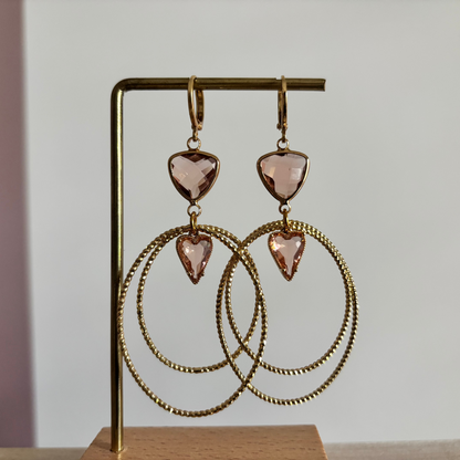 Symmetrische goudkleurige oorbellen Liene aan een rekje, opgebouwd uit een driehoekige champagnekleurige glasbedel, twee robuuste ovale ringen en een klein champagnekleurig hartje als verbinding.