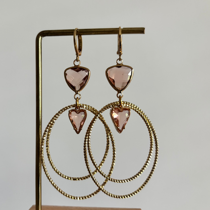 Symmetrische goudkleurige oorbellen Liene aan een rekje, opgebouwd uit een driehoekige champagnekleurige glasbedel, twee robuuste ovale ringen en een klein champagnekleurig hartje als verbinding.