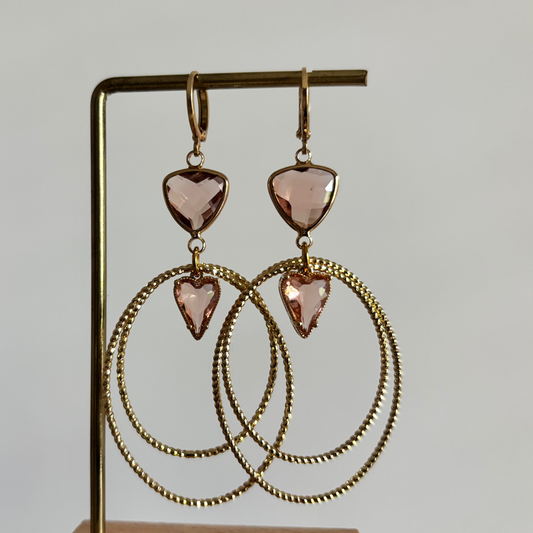 Symmetrische goudkleurige oorbellen Liene aan een rekje, opgebouwd uit een driehoekige champagnekleurige glasbedel, twee robuuste ovale ringen en een klein champagnekleurig hartje als verbinding.