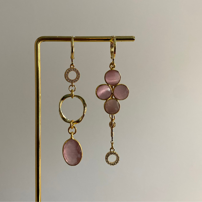 Asymmetrisch oorbellenpaar ‘Lola’: linker oorbel met zirkonia steentje, mini twisted ring en roze glazen bedel; rechter oorbel met taupekleurige steen, kwastje met sleuteltje en zirkonia steentje. Verguld in 14k goud, minimalistisch ontwerp met subtiele details.