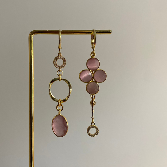 Asymmetrisch oorbellenpaar ‘Lola’: linker oorbel met zirkonia steentje, mini twisted ring en roze glazen bedel; rechter oorbel met taupekleurige steen, kwastje met sleuteltje en zirkonia steentje. Verguld in 14k goud, minimalistisch ontwerp met subtiele details.