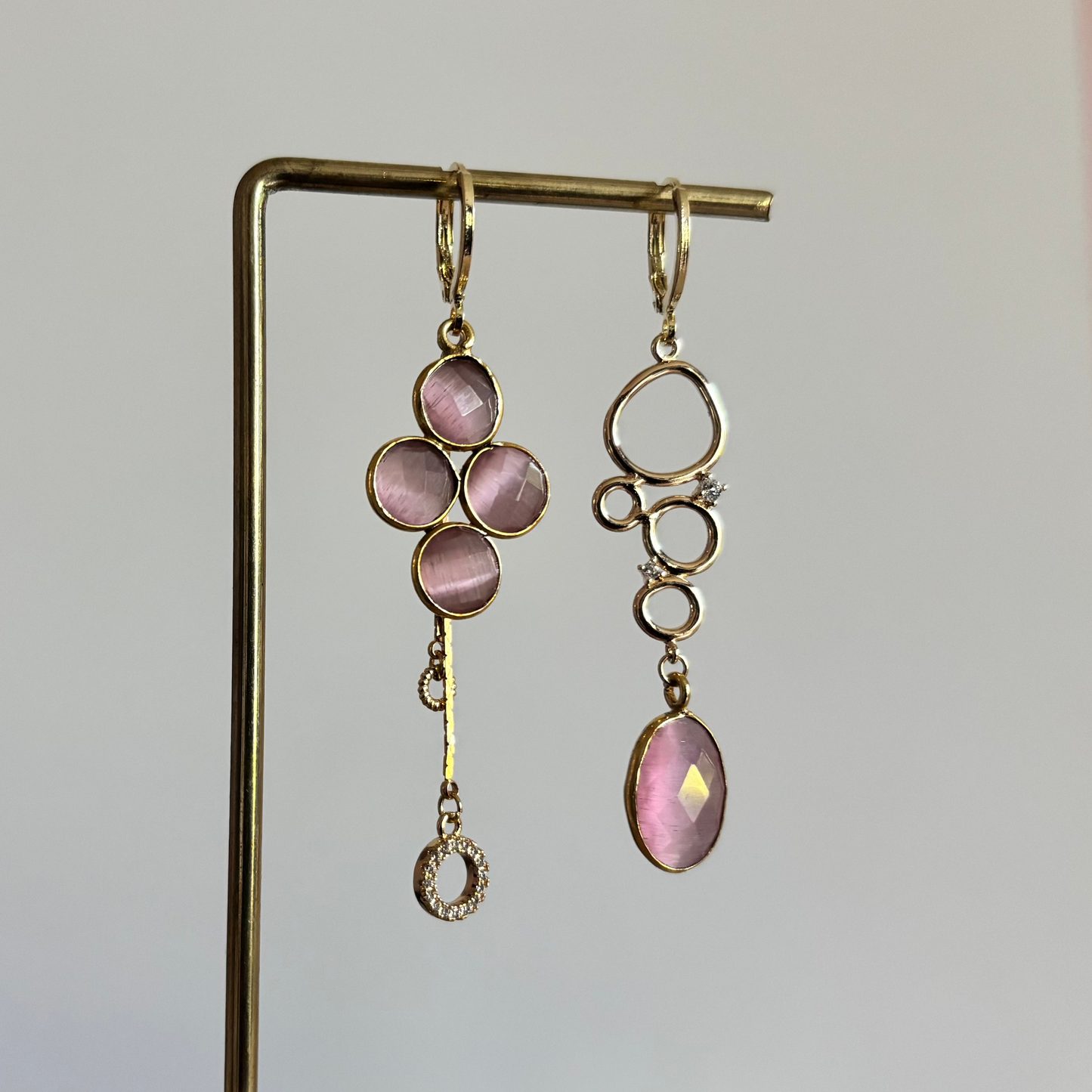 symmetrische 14k vergulde oorbellen met zachtroze stenen op een houten display. De linker oorbel heeft een klavervorm van roze stenen met een zirkonia ringetje; de rechter oorbel bestaat uit gouden cirkels met een druppelvormige roze steen.