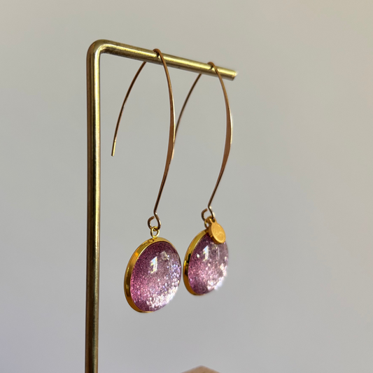 "Lange goudkleurige oorbellen met een ronde, glitter roze cabochon bedel van 18 mm hangend aan een display."