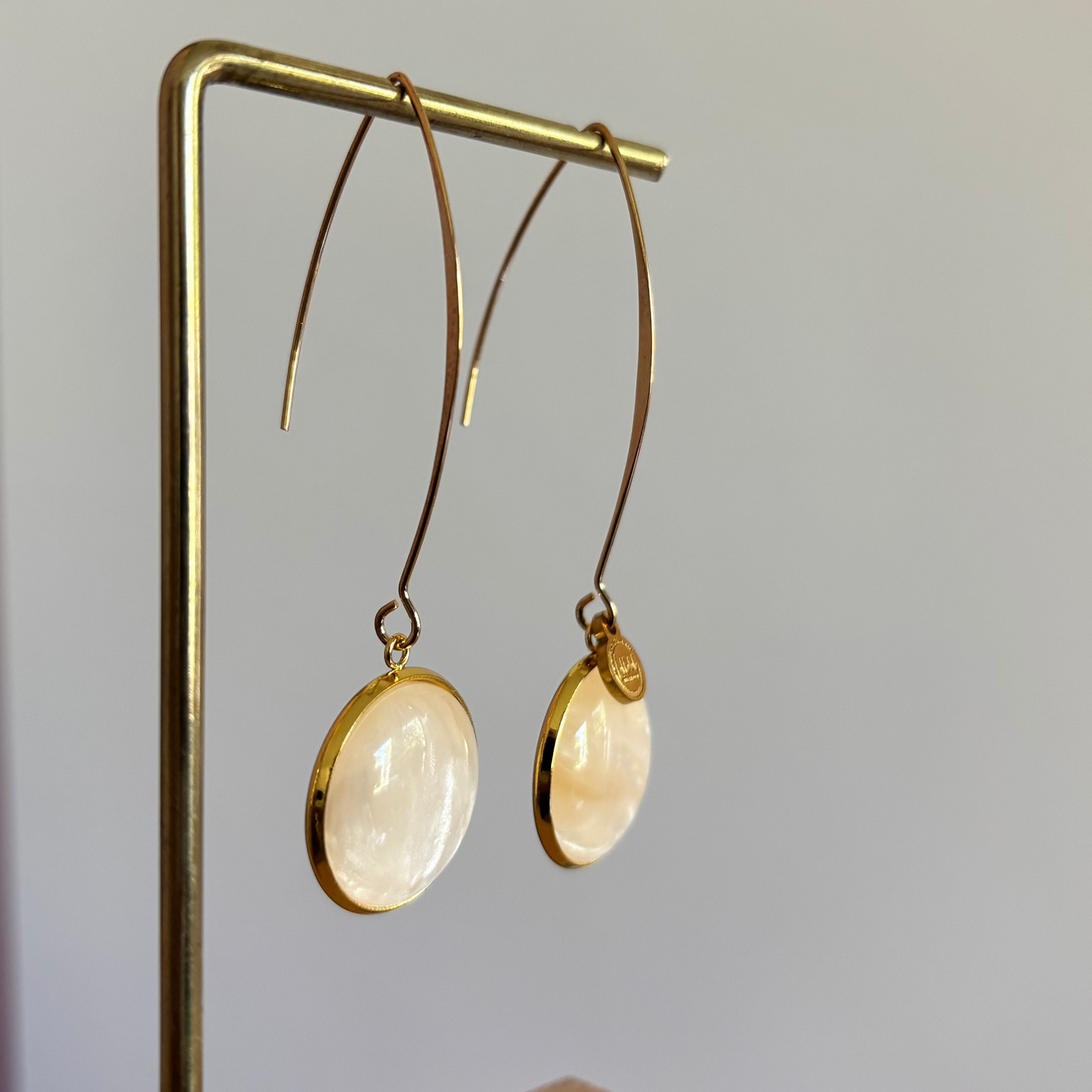"Lange goudkleurige oorbellen met een ronde, parelmoer cabochon bedel van 18 mm hangend aan een display."