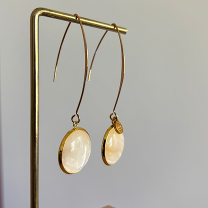 "Lange goudkleurige oorbellen met een ronde, parelmoer cabochon bedel van 18 mm hangend aan een display."