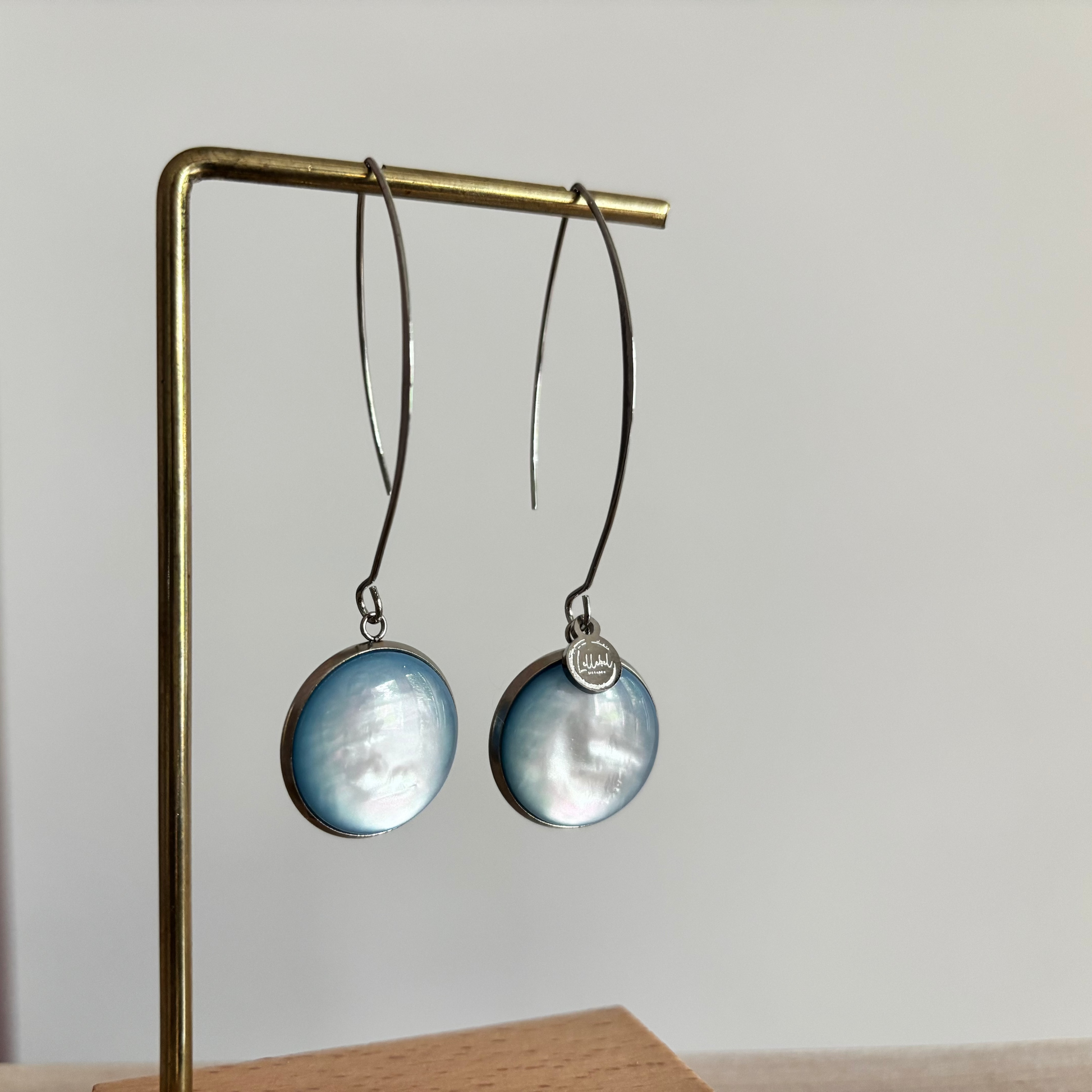 Symmetrische zilverkleurige oorbellen Lottie aan een rekje, voorzien van een lange steker, een grote ronde lichtblauwe cabochon kraal en een klein zilveren merktagje.