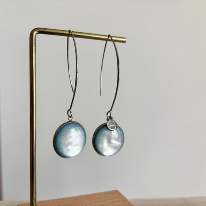 Symmetrische zilverkleurige oorbellen Lottie aan een rekje, voorzien van een lange steker, een grote ronde lichtblauwe cabochon kraal en een klein zilveren merktagje.