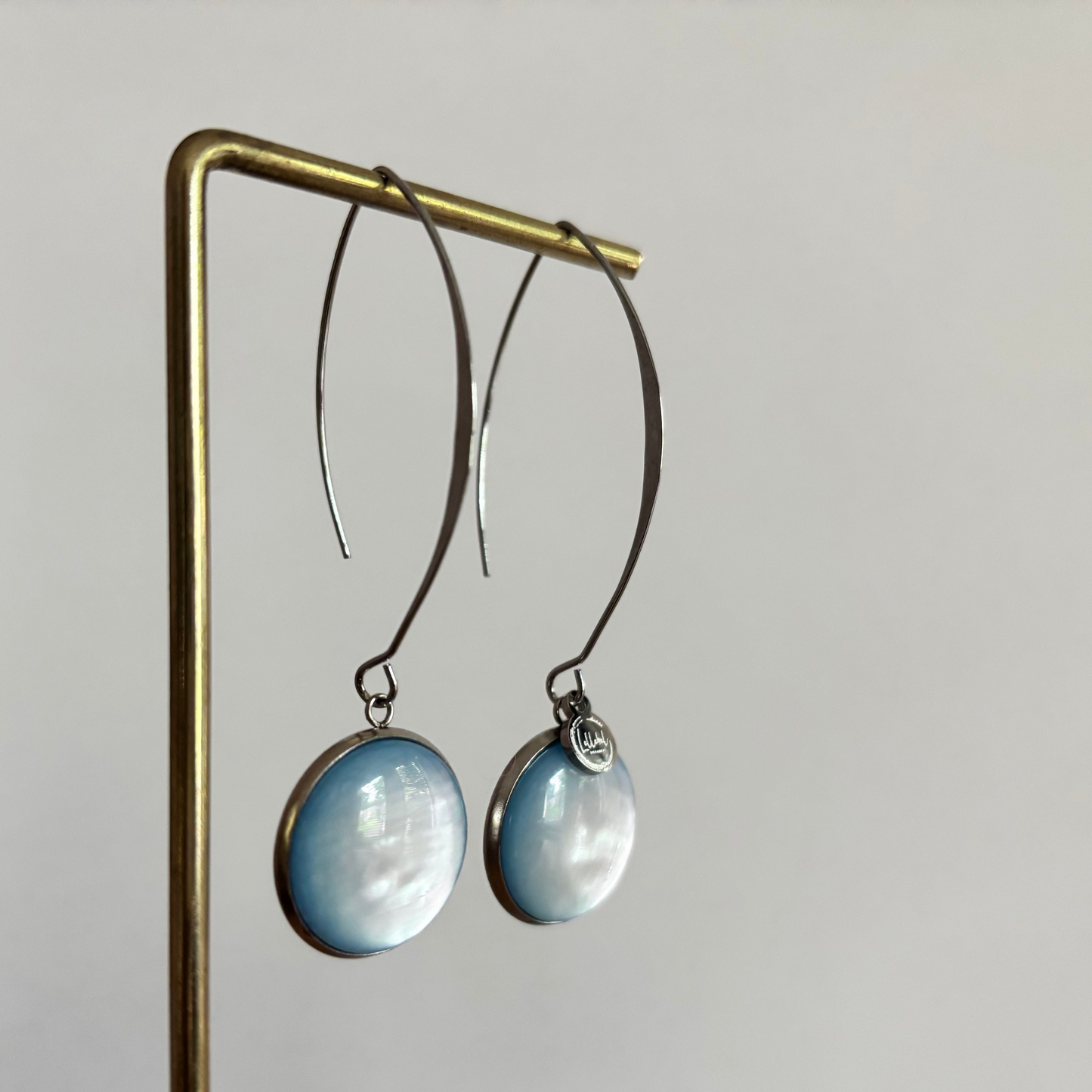 Symmetrische zilverkleurige oorbellen Lottie aan een rekje, voorzien van een lange steker, een grote ronde lichtblauwe cabochon kraal en een klein zilveren merktagje.