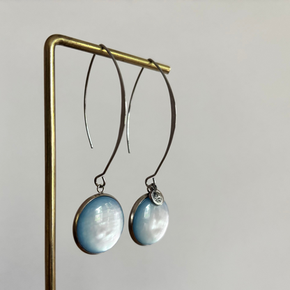 Symmetrische zilverkleurige oorbellen Lottie aan een rekje, voorzien van een lange steker, een grote ronde lichtblauwe cabochon kraal en een klein zilveren merktagje.