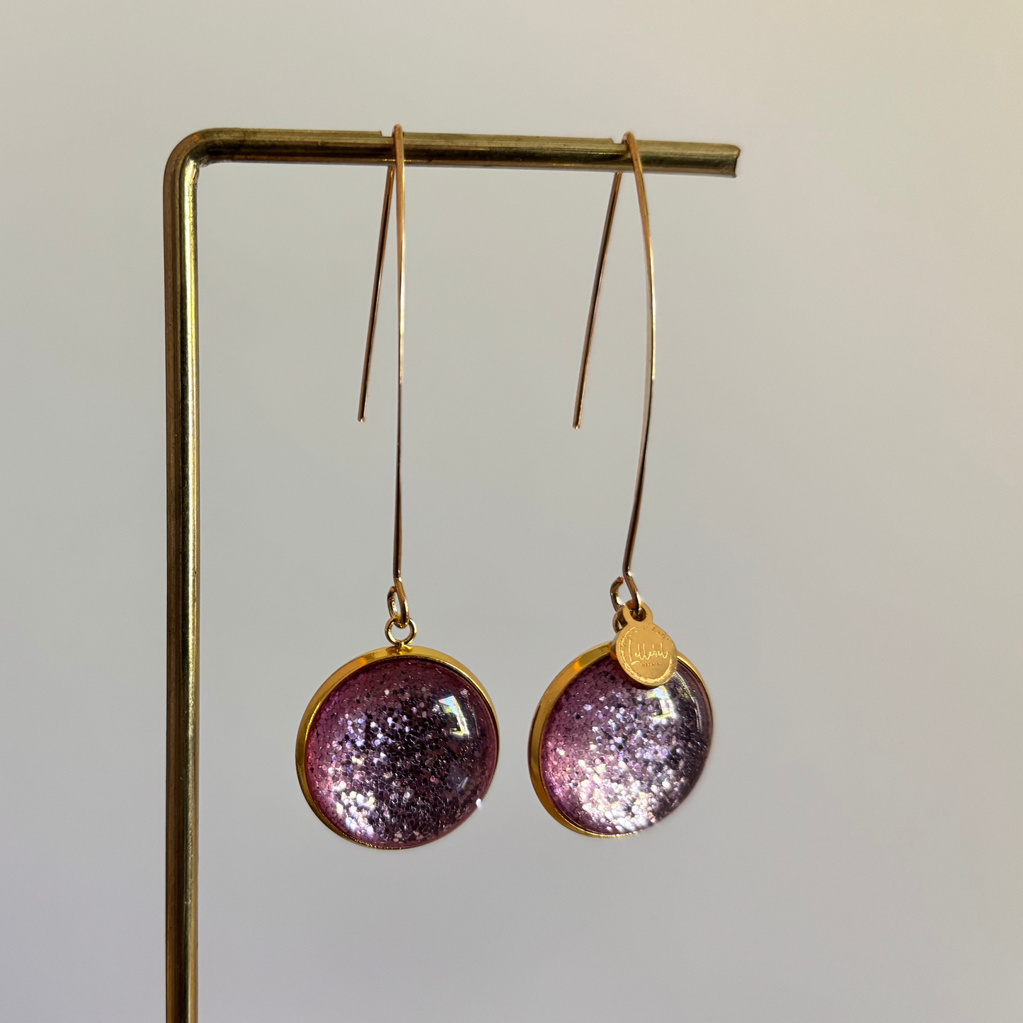 "Lange goudkleurige oorbellen met een ronde, glitter roze cabochon bedel van 18 mm hangend aan een display."