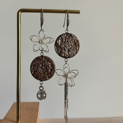 Asymmetrische zilverkleurige oorbellen Lou aan een rekje. Links een open zilveren draadbloem met een ronde bronsbruine glittersteen en een ronde zirkonia hanger; rechts een bronsbruine glittersteen met een draadbloem en een lang kettinkje.
