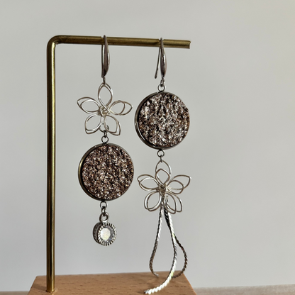 Asymmetrische zilverkleurige oorbellen Lou aan een rekje. Links een open zilveren draadbloem met een ronde bronsbruine glittersteen en een ronde zirkonia hanger; rechts een bronsbruine glittersteen met een draadbloem en een lang kettinkje.