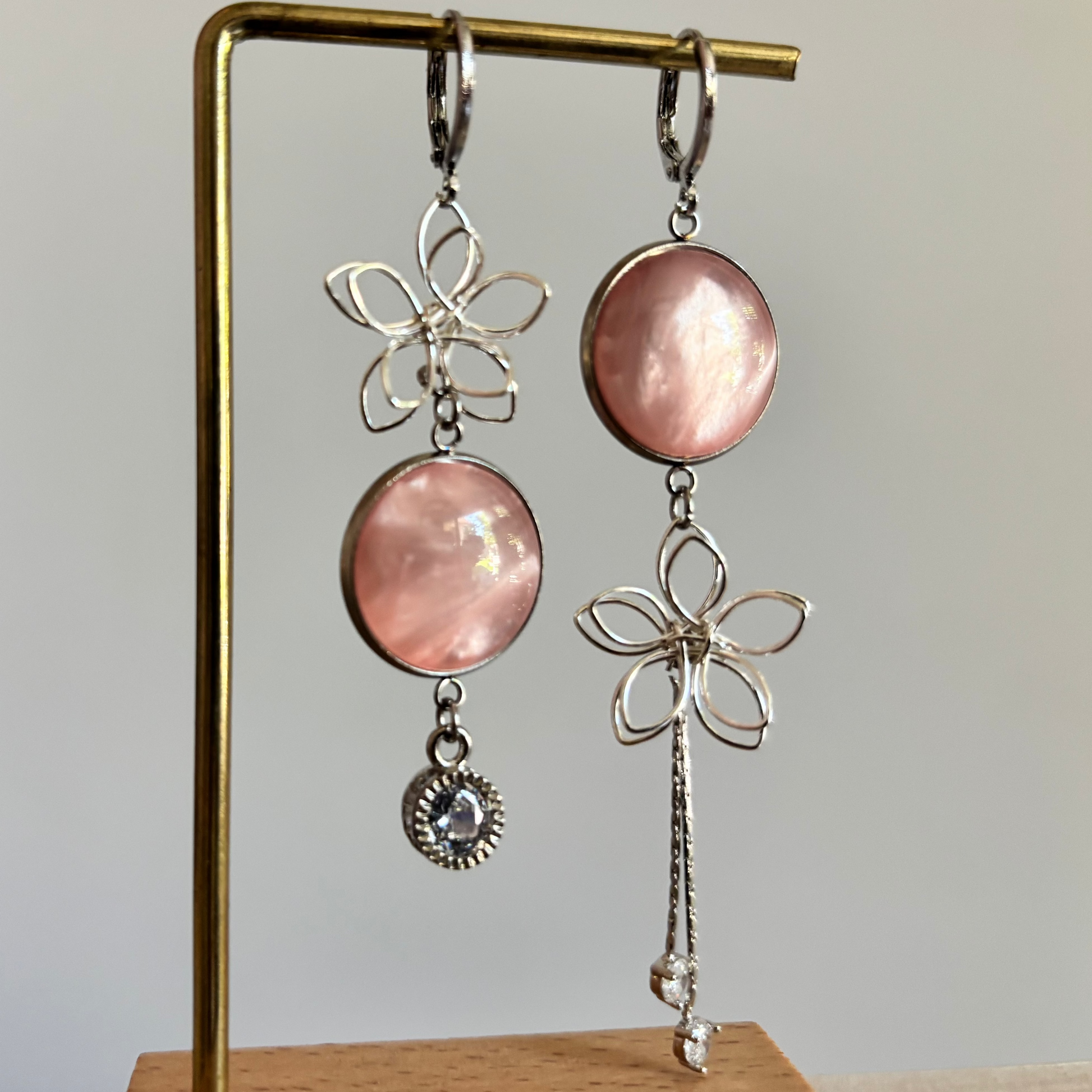 Asymmetrische zilveren oorbellen van Lellebel op een gouden standaard. De oorbellen bestaan uit een combinatie van zilveren draadbloemen, ronde zalmroze glansbedels, een klein zirkonia steentje aan de ene kant en een langer zilveren kettinkje aan de andere kant.