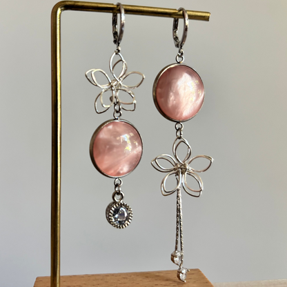 Asymmetrische zilveren oorbellen van Lellebel op een gouden standaard. De oorbellen bestaan uit een combinatie van zilveren draadbloemen, ronde zalmroze glansbedels, een klein zirkonia steentje aan de ene kant en een langer zilveren kettinkje aan de andere kant.