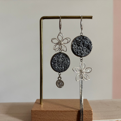 Asymmetrische zilverkleurige oorbellen Lou aan een rekje. Links een open zilveren draadbloem met een ronde lichtblauwe glittersteen en een ronde zirkonia hanger; rechts een lichtblauweglittersteen met een draadbloem en een lang kettinkje.