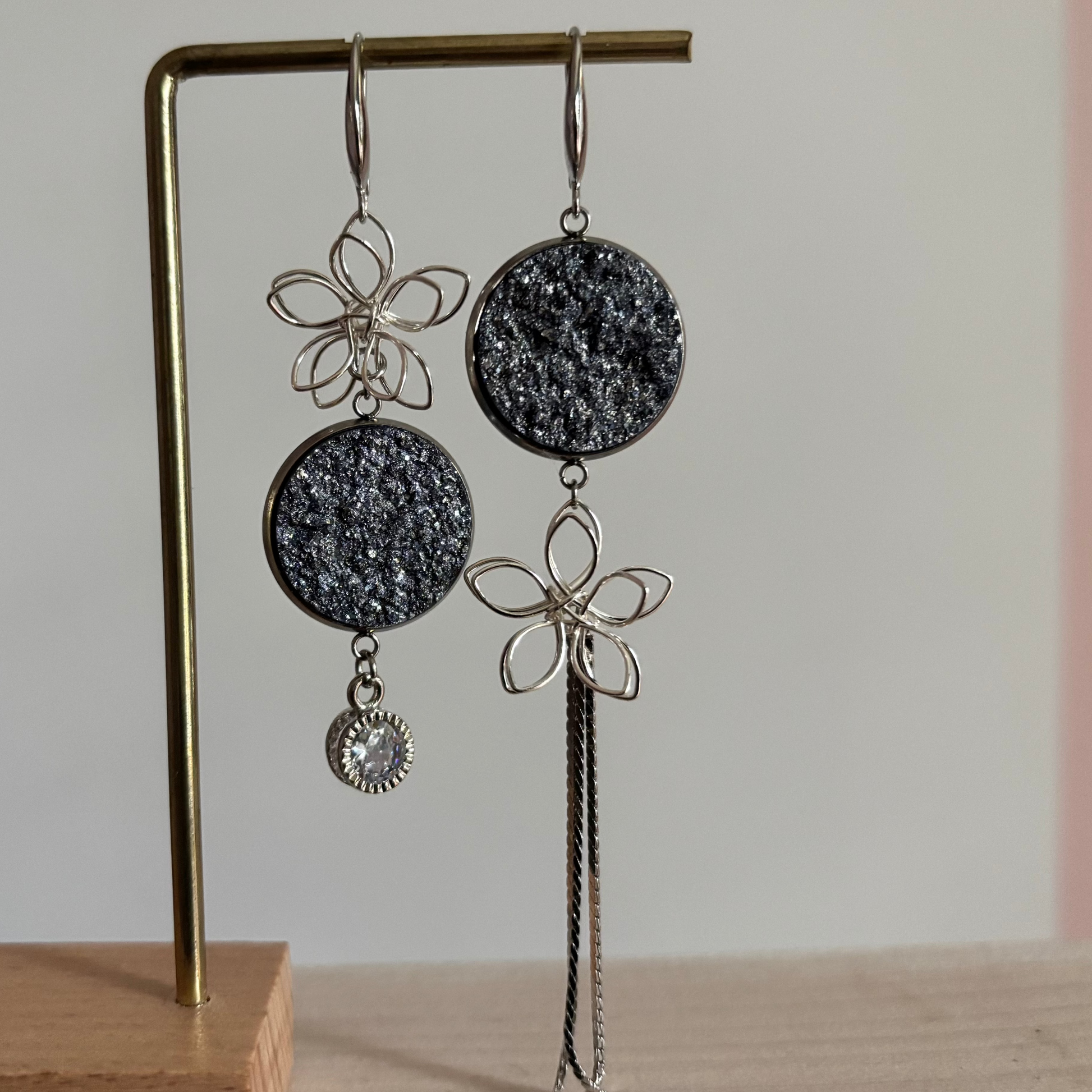 Asymmetrische zilverkleurige oorbellen Lou aan een rekje. Links een open zilveren draadbloem met een ronde lichtblauwe glittersteen en een ronde zirkonia hanger; rechts een lichtblauweglittersteen met een draadbloem en een lang kettinkje.