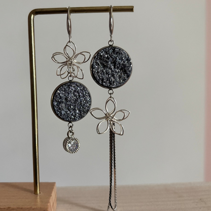 Asymmetrische zilverkleurige oorbellen Lou aan een rekje. Links een open zilveren draadbloem met een ronde lichtblauwe glittersteen en een ronde zirkonia hanger; rechts een lichtblauweglittersteen met een draadbloem en een lang kettinkje.