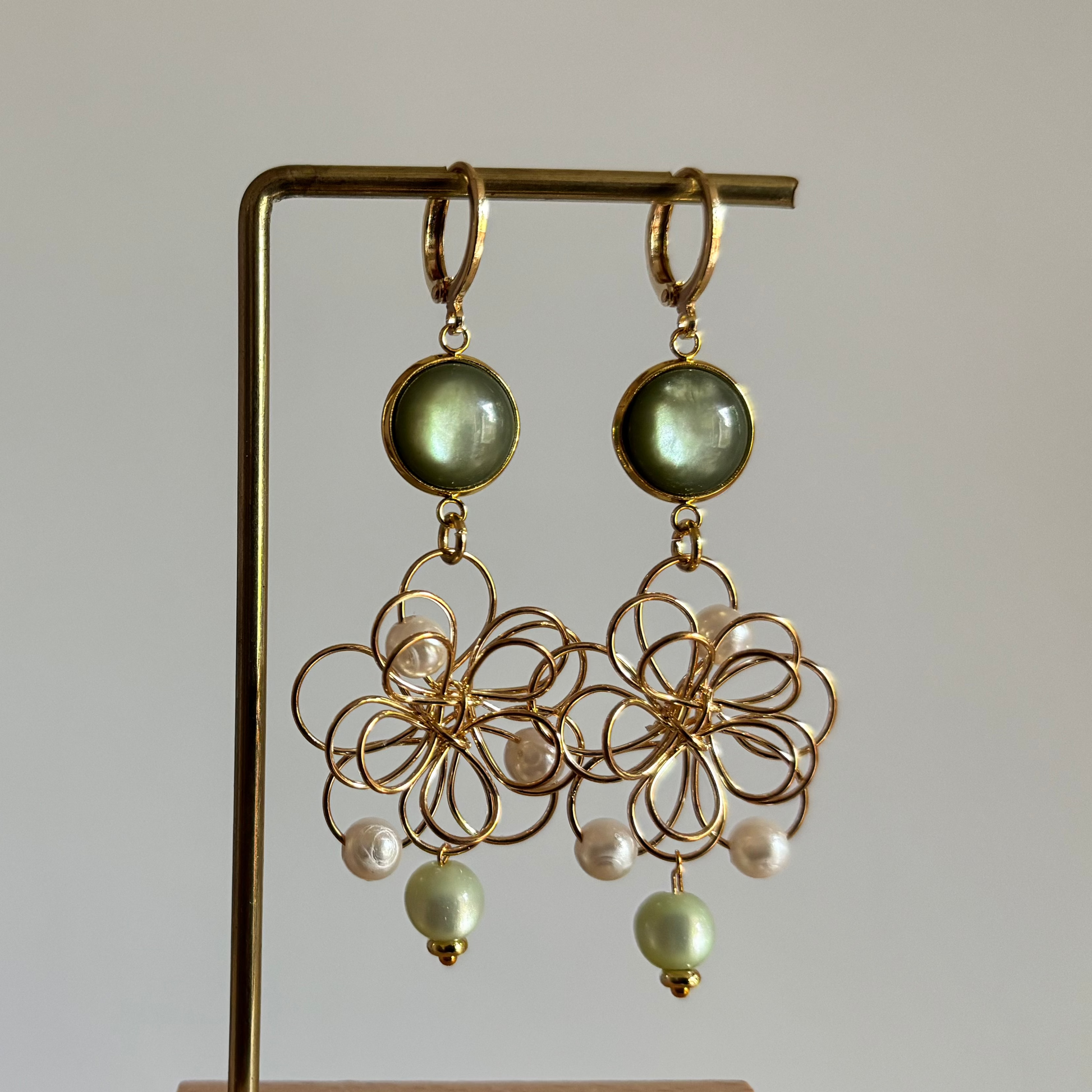 Symmetrische goudkleurige oorbellen 'Louise Groen' op een houten display. De oorbellen bestaan uit een ronde olijfgroene steen en een grote opengewerkte bloemhanger van goudkleurig draad met witte en lichtgroene pareltjes.