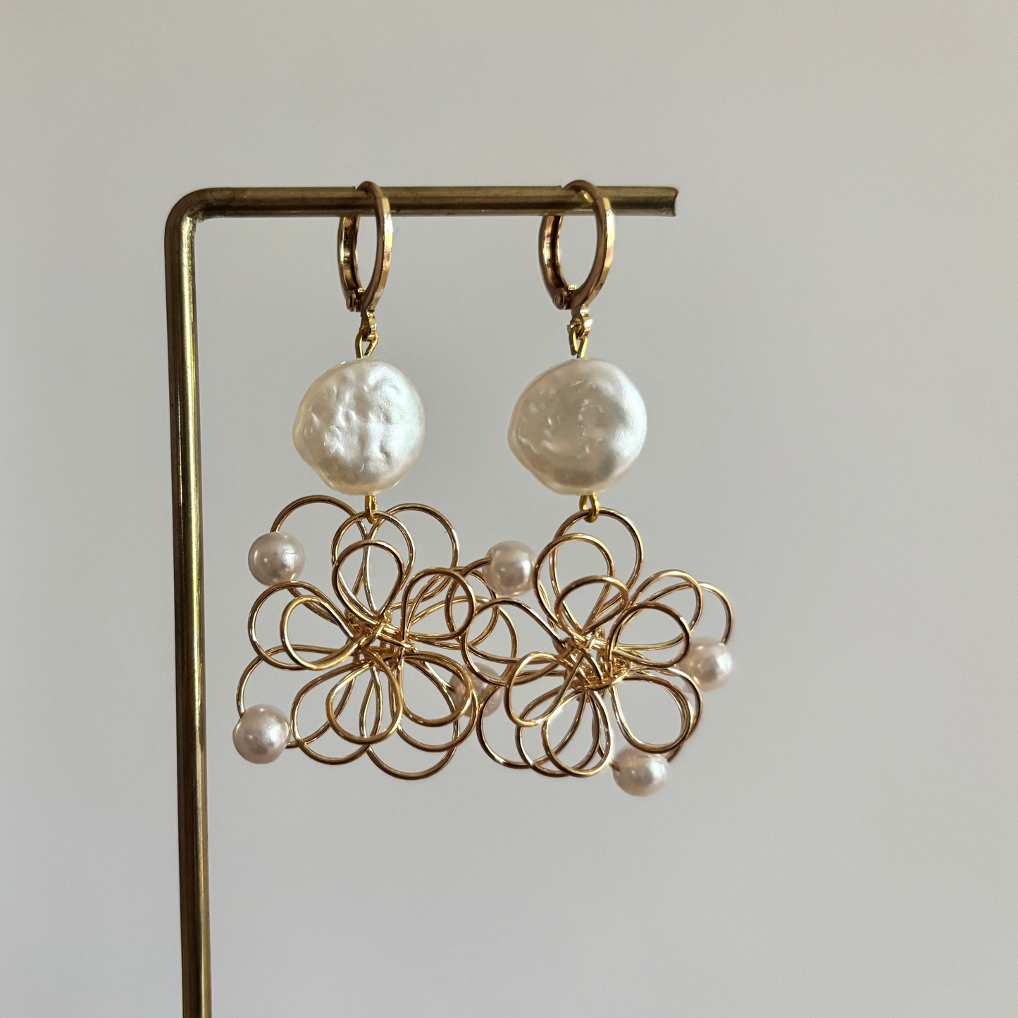 Symmetrische goudkleurige oorbellen 'Louise Parelmoer' op een houten display. De oorbellen bestaan uit een ronde, glanzende witte parelmoersteen en een grote opengewerkte bloemhanger van goudkleurig draad met kleine witte pareltjes.
