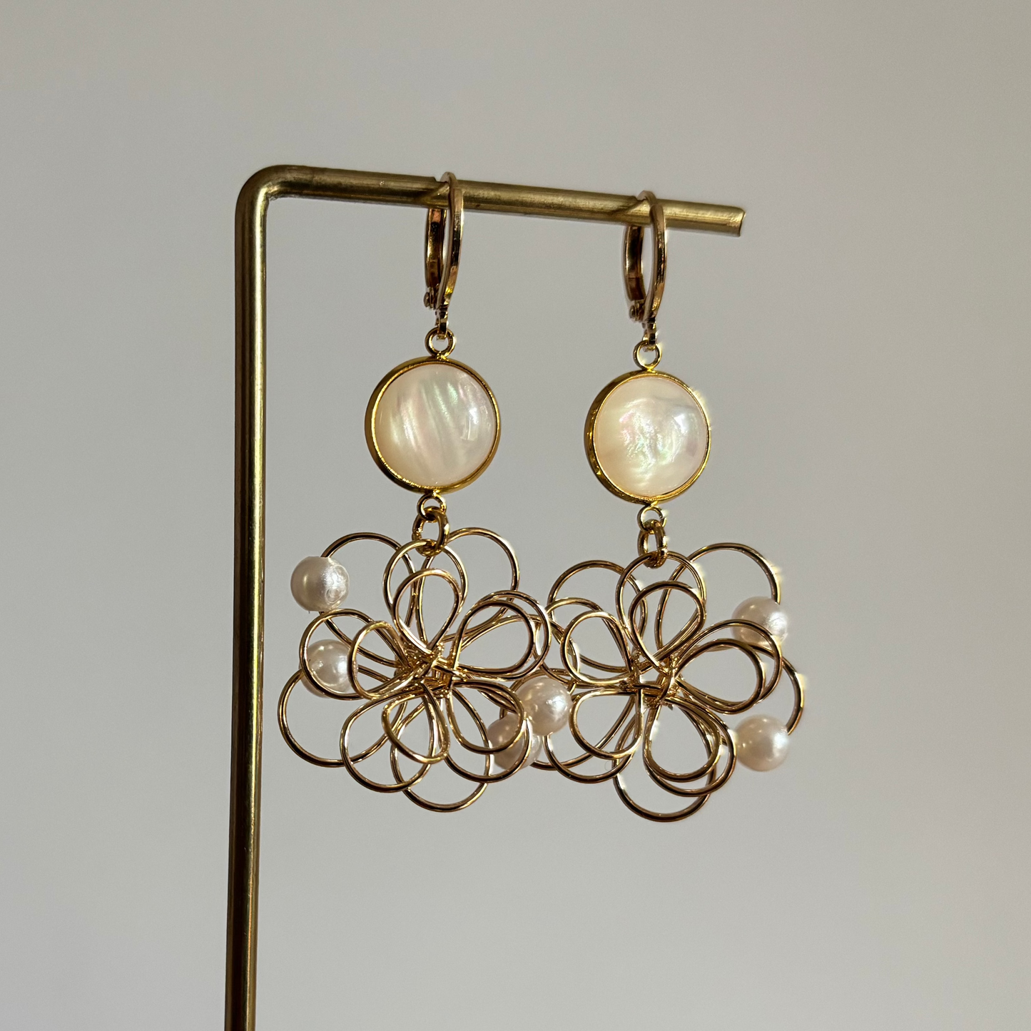 Symmetrische goudkleurige oorbellen 'Louise Parelmoer' op een houten display. De oorbellen bestaan uit een ronde, glanzende witte parelmoersteen en een grote opengewerkte bloemhanger van goudkleurig draad met kleine witte pareltjes.