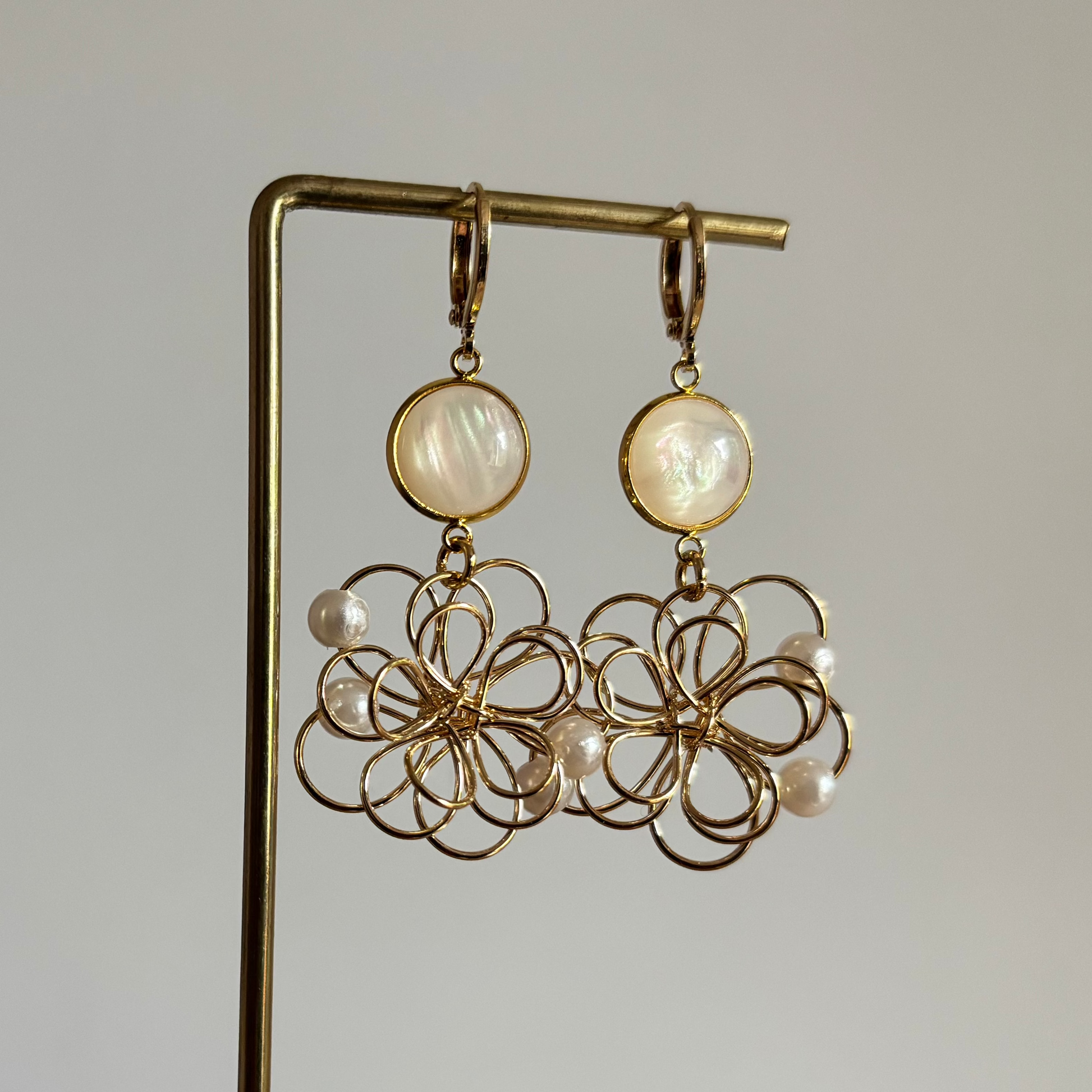 Symmetrische goudkleurige oorbellen 'Louise Parelmoer' op een houten display. De oorbellen bestaan uit een ronde, glanzende witte parelmoersteen en een grote opengewerkte bloemhanger van goudkleurig draad met kleine witte pareltjes.