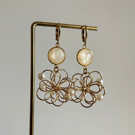 Symmetrische goudkleurige oorbellen 'Louise Parelmoer' op een houten display. De oorbellen bestaan uit een ronde, glanzende witte parelmoersteen en een grote opengewerkte bloemhanger van goudkleurig draad met kleine witte pareltjes.