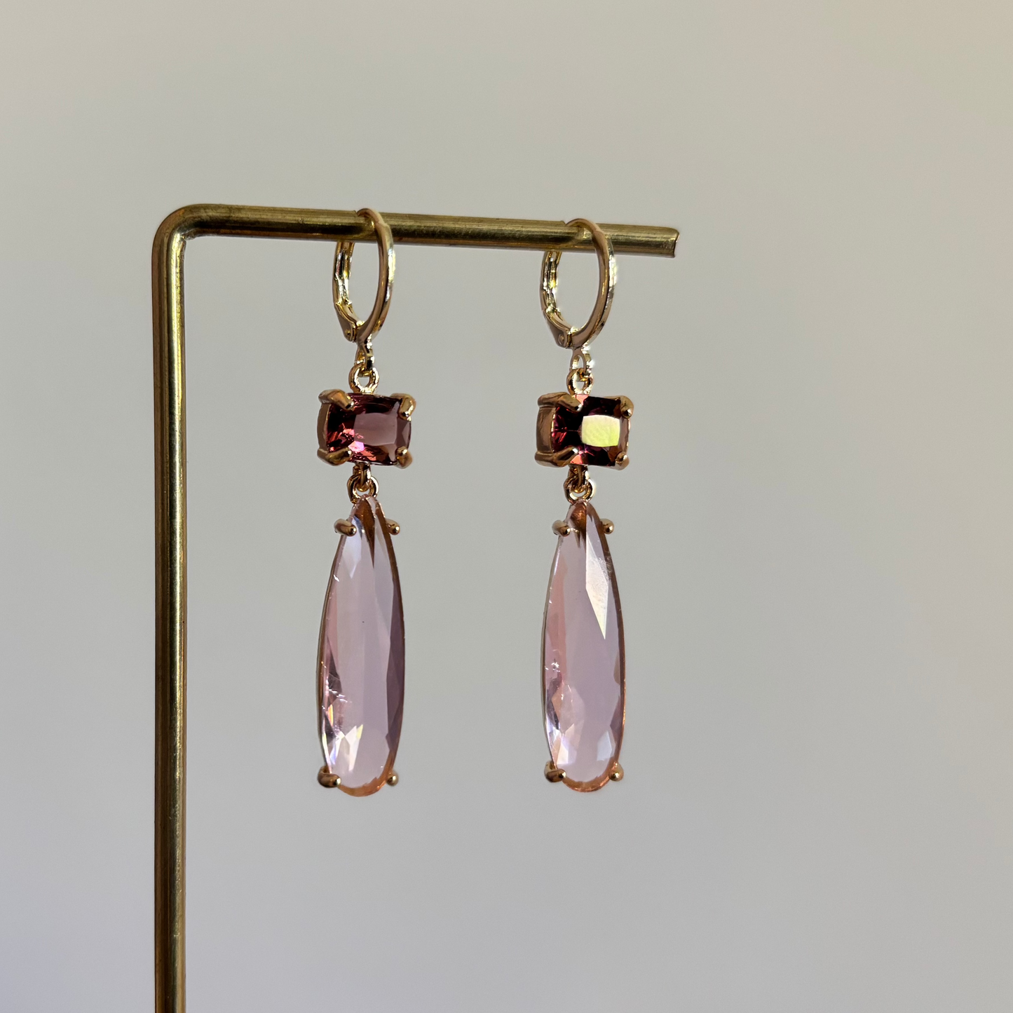 Goudkleurige oorbellen Maya met een kleine vierkante burgundy steen en een lange lichtroze druppelvormige hanger van kristal.