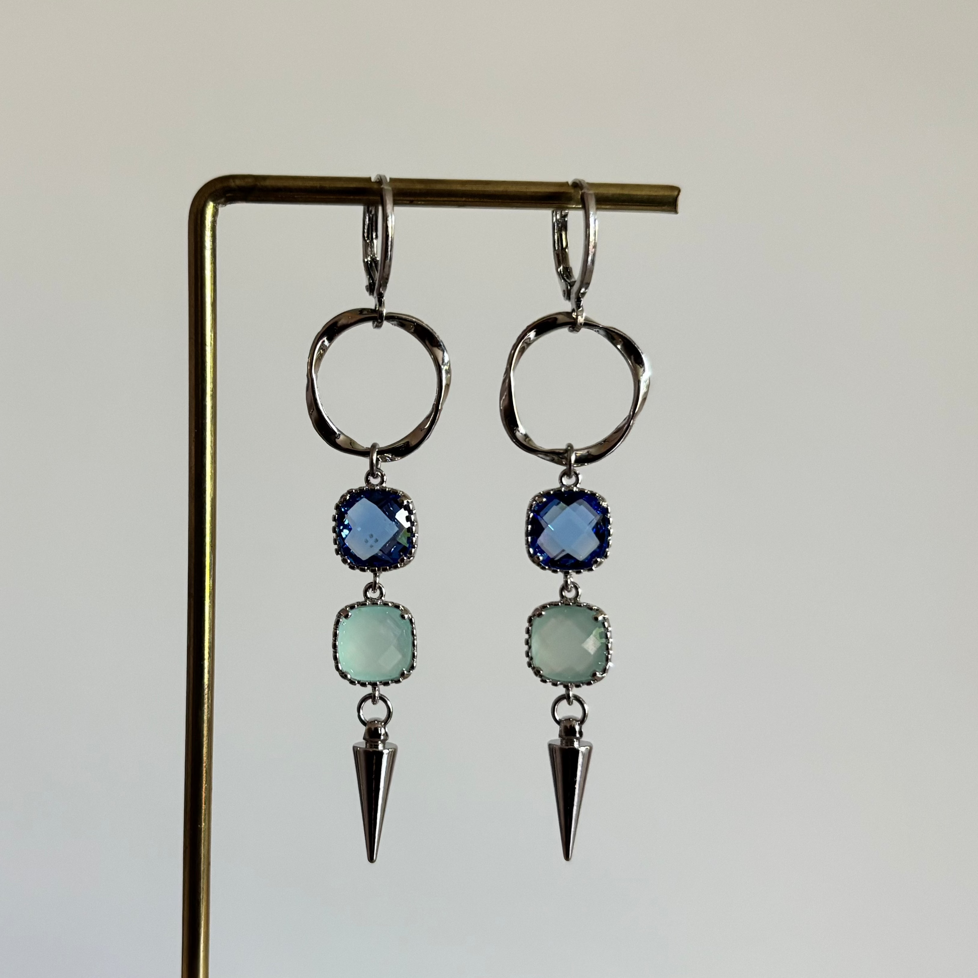 Zilverkleurige oorbellen 'Melly Blauw' op een houten display. De oorbellen bestaan uit een gedraaide cirkel, een donkerblauwe steen, een lichtblauwe steen en een zilveren kegel.