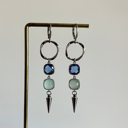 Zilverkleurige oorbellen 'Melly Blauw' op een houten display. De oorbellen bestaan uit een gedraaide cirkel, een donkerblauwe steen, een lichtblauwe steen en een zilveren kegel.