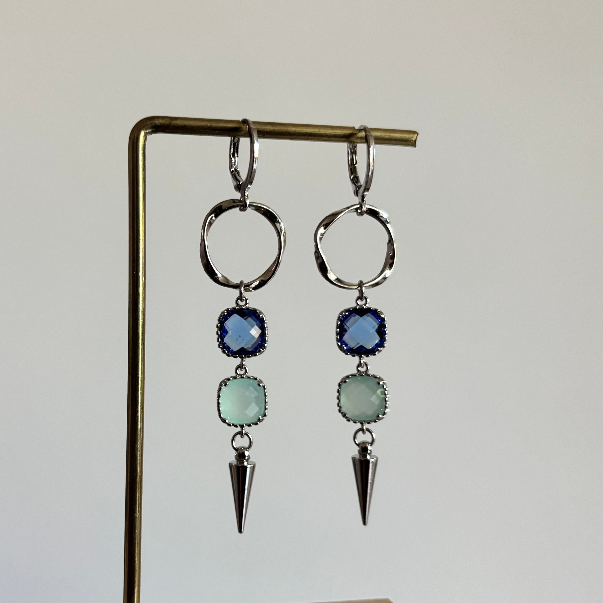 Zilverkleurige oorbellen 'Melly Blauw' op een houten display. De oorbellen bestaan uit een gedraaide cirkel, een donkerblauwe steen, een lichtblauwe steen en een zilveren kegel.