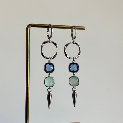 Zilverkleurige oorbellen 'Melly Blauw' op een houten display. De oorbellen bestaan uit een gedraaide cirkel, een donkerblauwe steen, een lichtblauwe steen en een zilveren kegel.